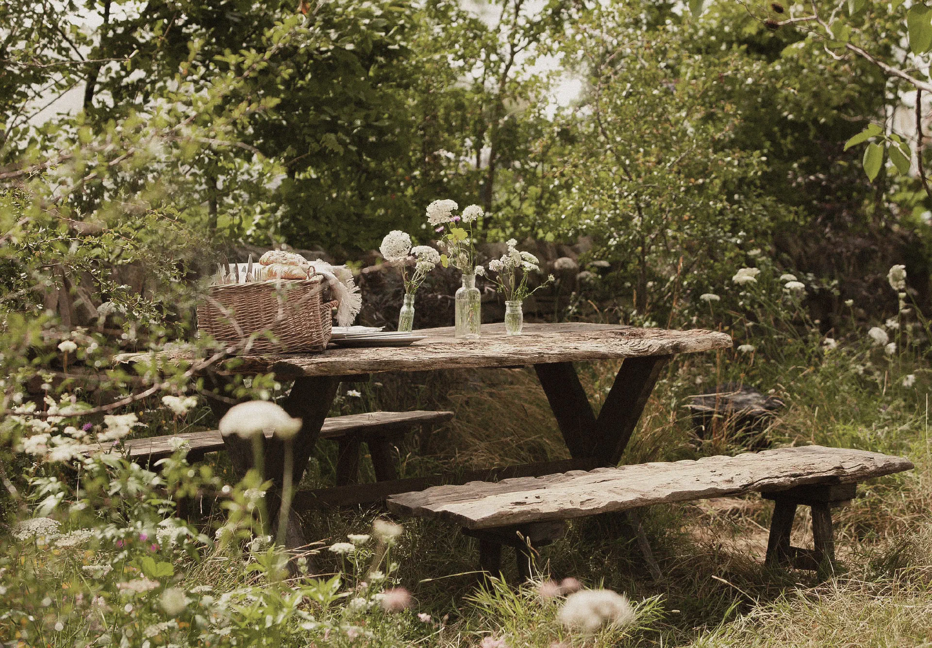 Rustic timber picnic table laid with a wicker basket and jars of wildflowers, in long meadow grass